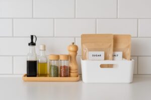 白いキッチンカウンターの上に整然と配置された小さな調味料ボトルと保存容器。ラベル付きでカテゴリ別に分けられ、清潔感のある収納が印象的。