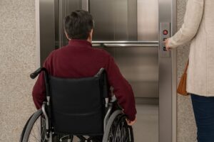 車いす利用者がエレベーターに入る場面で、介助者が「開」ボタンを押してサポートしている様子。
