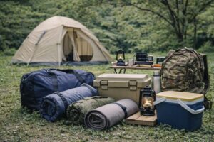キャンプ場のテント横に、1泊分の着替えや寝具、収納ケースなどの荷物がまとめて置かれている様子。宿泊キャンプの準備をイメージした風景。