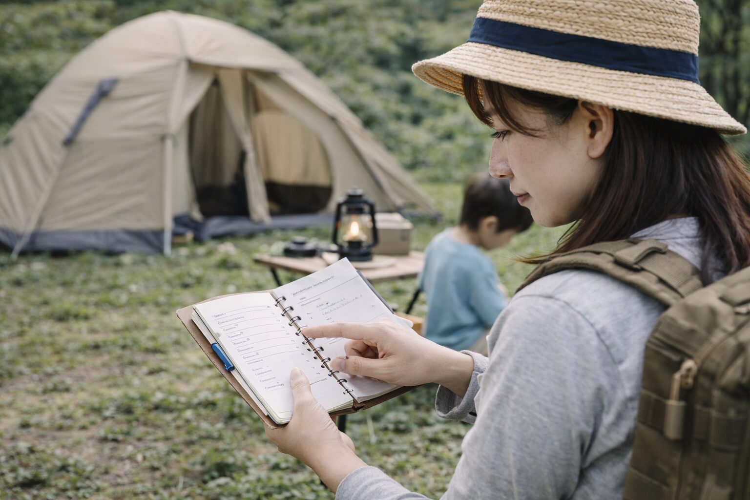 キャンプ場で母親がチェックリストやスマートフォンを見ながら、当日の流れを確認している様子。周囲の環境を見渡しながら準備を進めている場面。